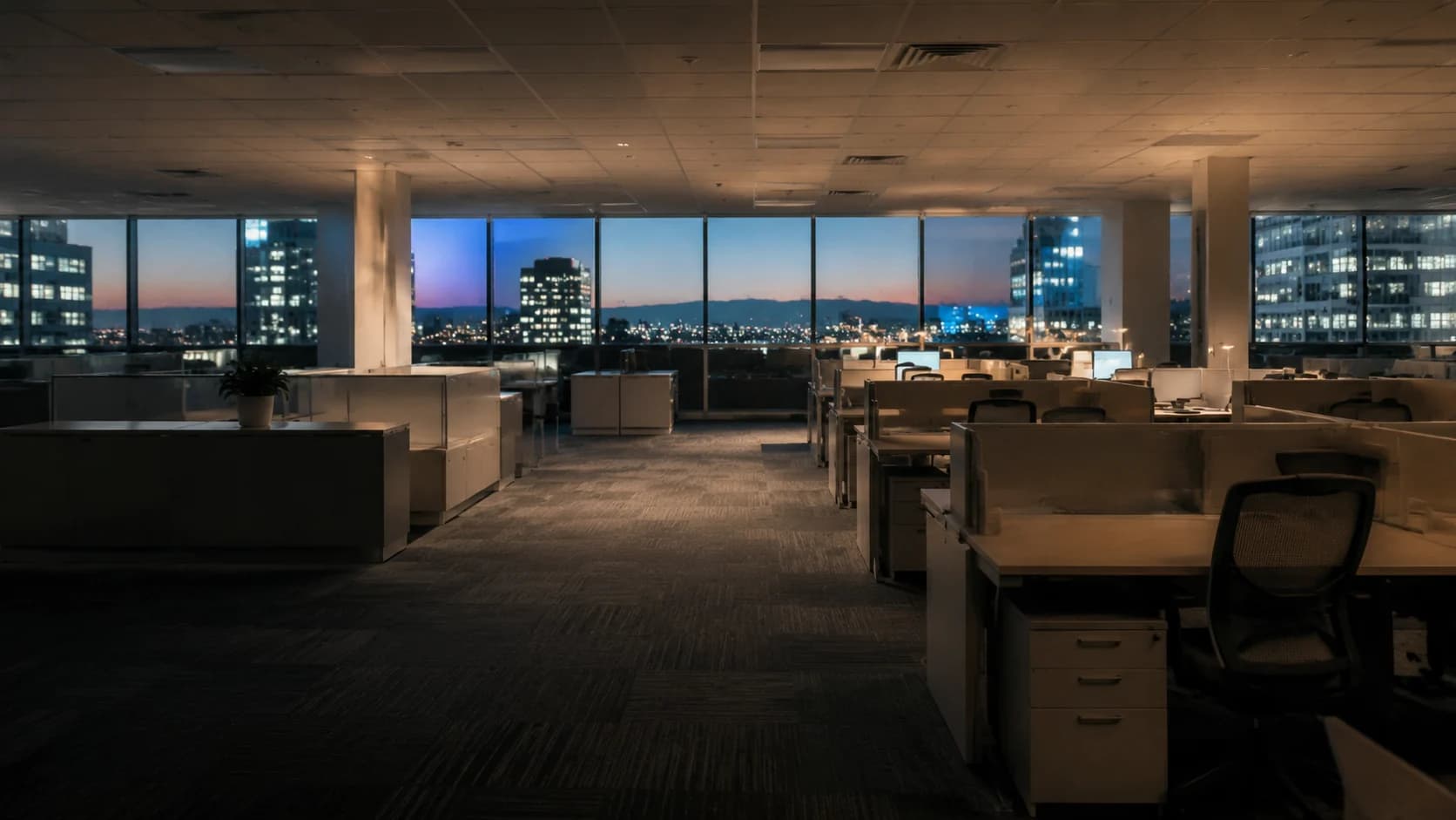 Half-empty office floorplate at dusk, vacant desks and a city skyline through the windows